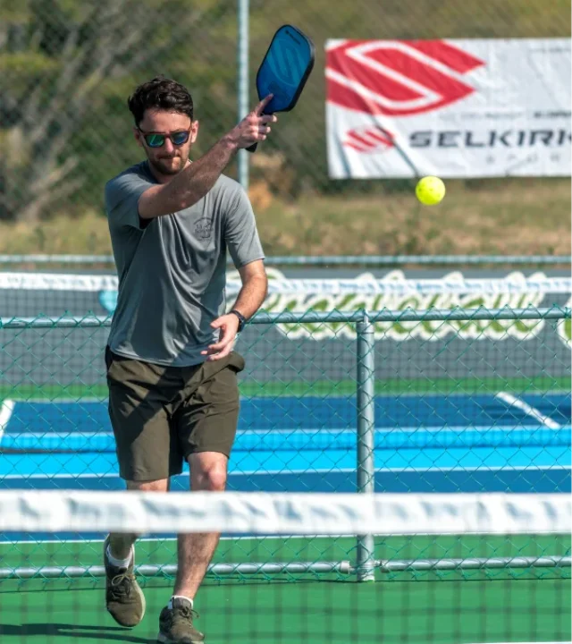 Young player wearing sunglasses hitting a yellow ball with a high overhead smash at Pickleball Club